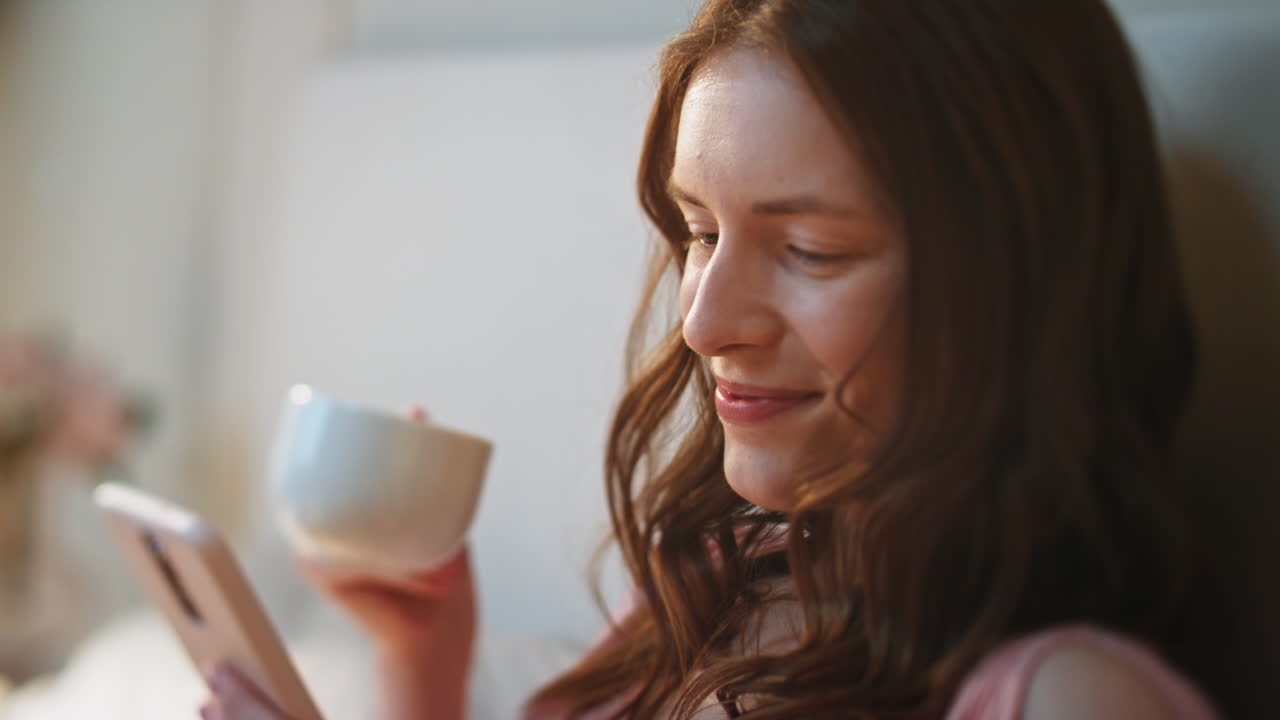 아침 클로즈업에서 커피를 즐기는 미소 짓는 여성. 휴대전화를 사용하는 평온한 여성