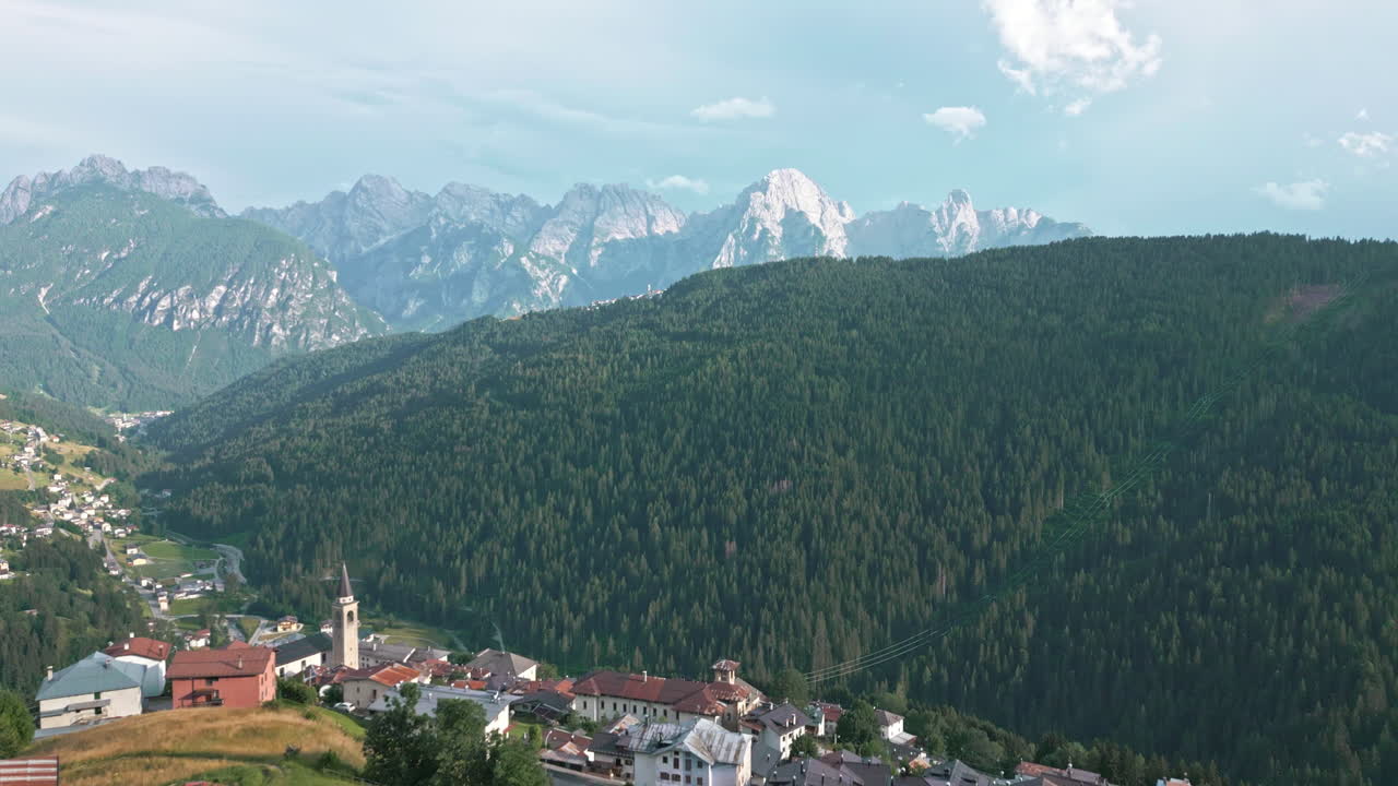 Drone video flying by historical hillside catholic church in old town surrounded by farms in the Dolomites Italy with mountain range in the background at sunset golden hour in summer