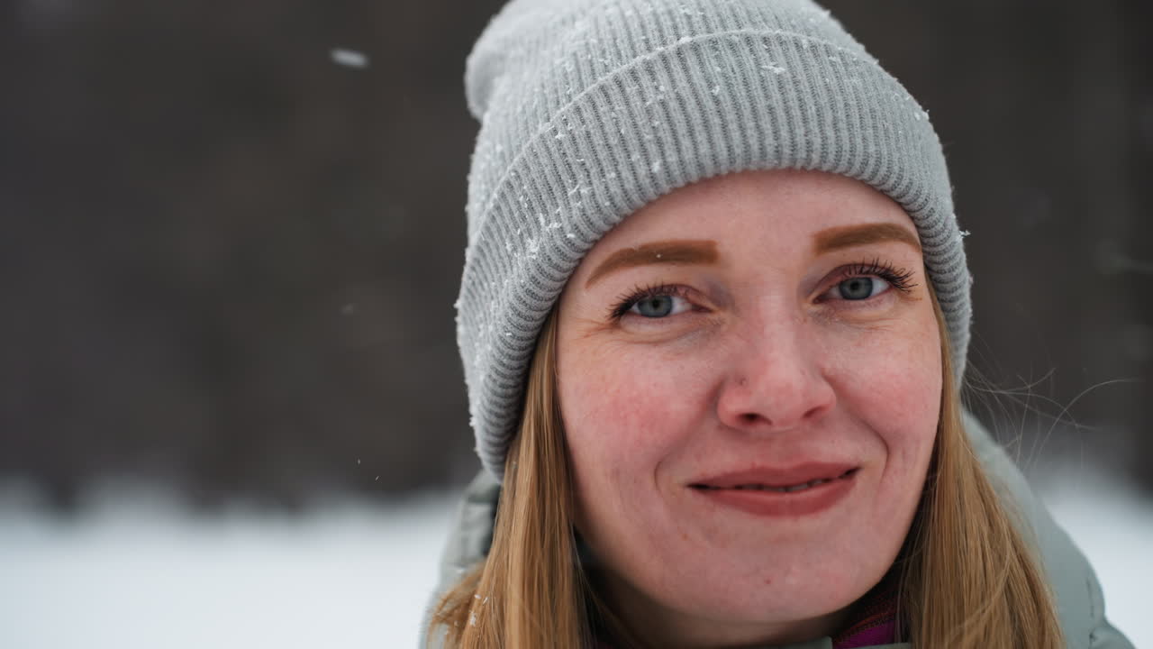 Woman wearing gray knit beanie and black gloves adjusting headwear during snowfall in snowy forest, smiling warmly while standing outside in winter clothing surrounded by falling snow and blurred trees