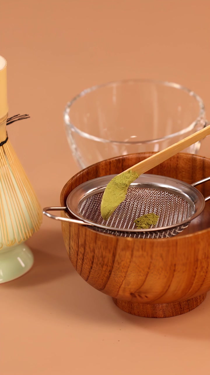 Primo piano di polvere di matcha versata con un cucchiaio in un setaccio di metallo sopra una ciotola di legno, con illuminazione da studio minimale.
