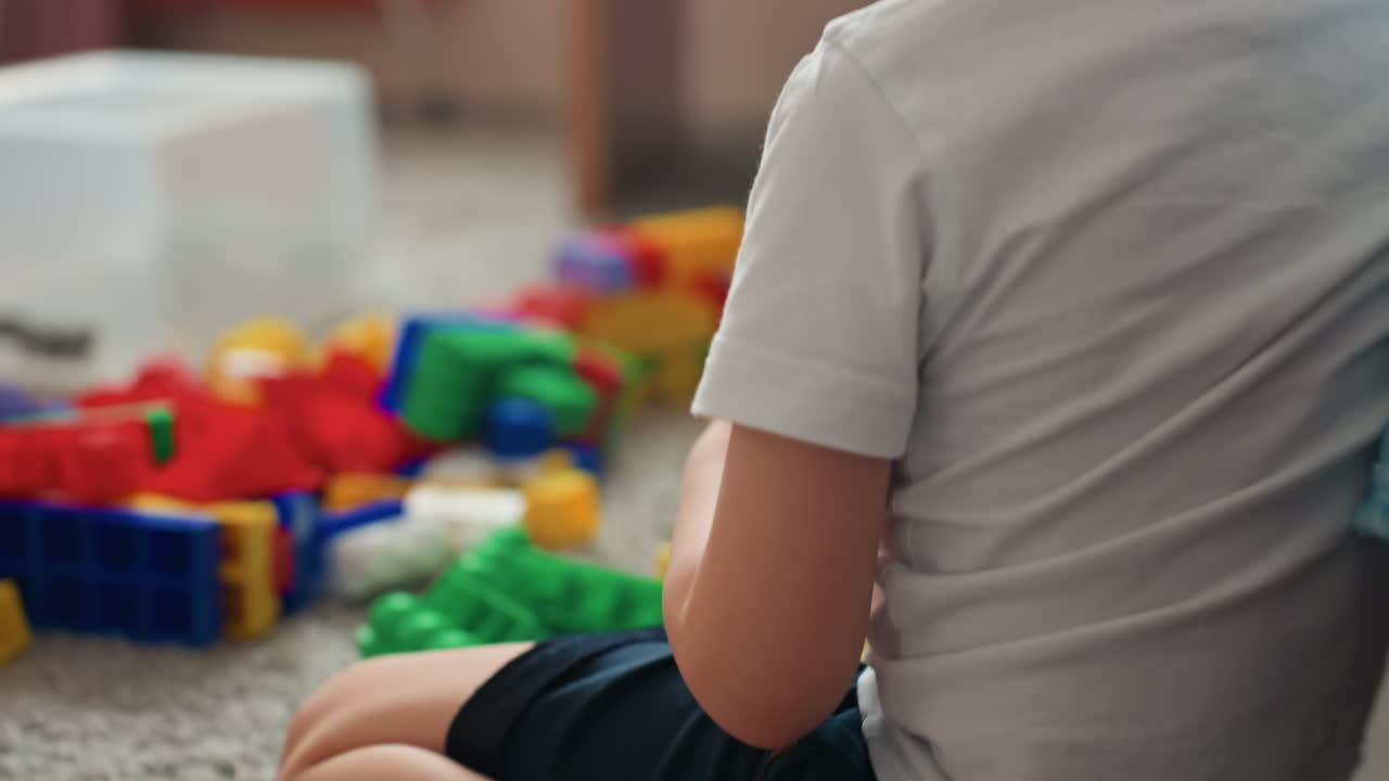 Un bambino seduto tra blocchi colorati sul tappeto, profilo pensieroso accanto a una coperta a righe. Tranquilla scena di camera da letto interna, primo piano di schiena e testa, una pausa contemplativa durante il gioco, atmosfera incentrata sul bambino.