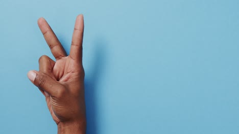 Close-up-of-hand-of-biracial-man-showing-peace-sign-with-copy-space-on-blue-background
