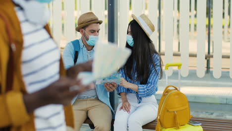 caucasian young happy man and woman travellers in facial masks sitting at bus stop talking and watching a map to plan a route