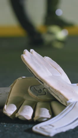 primer plano de guantes blancos de portero descansando cerca del poste de la portería en el césped con hojas secas esparcidas, fondo borroso de un jugador de fútbol equilibrando la pelota bajo los pies iluminado por las luces urbanas por la noche