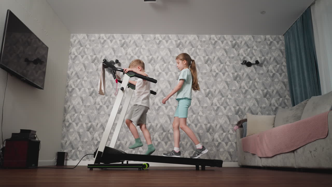 Two young siblings playing on treadmill in bright indoor room with patterned wall, wooden floor, and cozy sofa, boy leaning on handles while girl stands nearby observing and preparing to step on