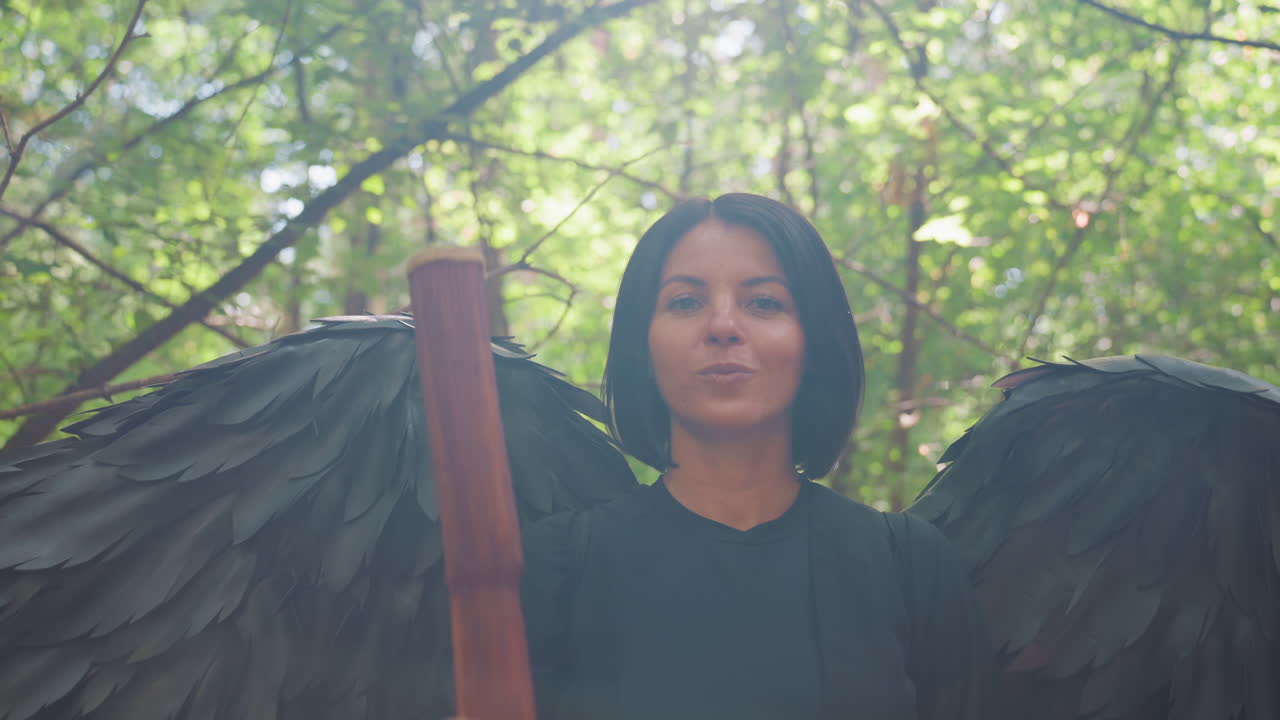 Young woman dressed in black wings rotates staff gracefully in green forest with sunlight shining from behind, creating cinematic fantasy scene filled with motion
