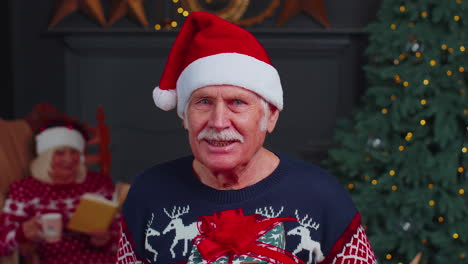 Senior-grandfather-in-festive-sweater-presenting-Christmas-gift-box,-smiling,-looking-at-camera