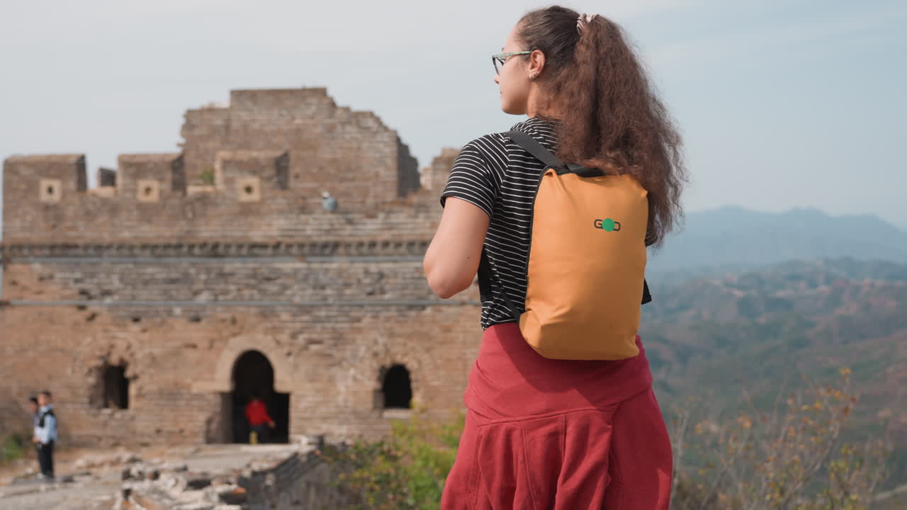 Donna backpacker in un'antica fortezza che domina la valle, travel blogger si ferma con zaino giallo e gonna rossa, cielo calmo e montagne lontane, archi in pietra e sentiero per visitatori, sincera contemplativa.