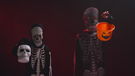 fotografía de estudio de niños vestidos con trajes de esqueleto haciendo trucos con cubos de dulces en halloween contra la luz de fondo de humo rojo
