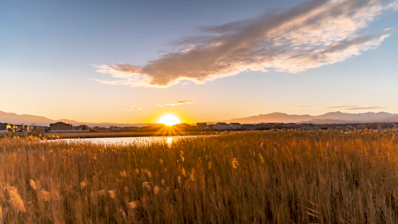 Sunset across a pond with pampas grass a wispy, colorful clouds crossing the sky with a suburb and mountains in the background - static time lapse
