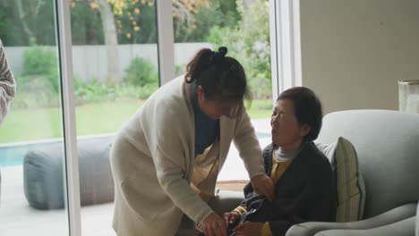 happy senior asian mother being made comfortable in armchair by adult daughter in living room