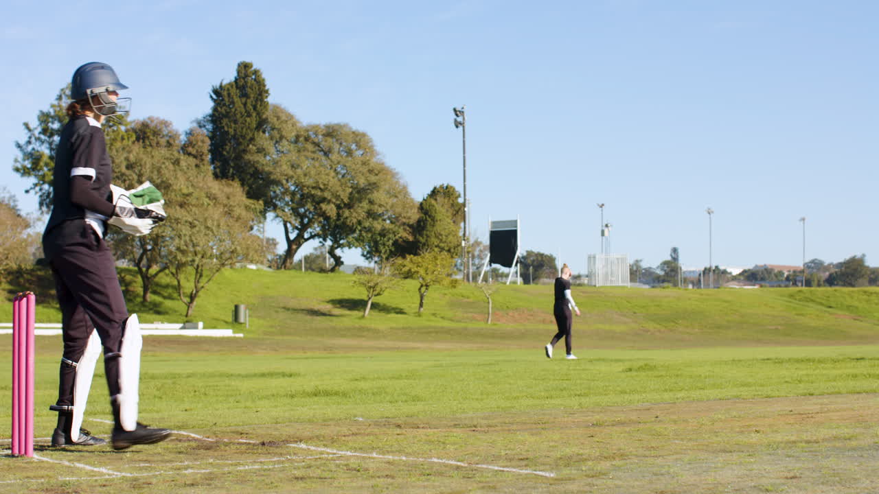 Playing cricket, woman in protective gear batting on field with bowler in distance
