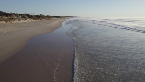 Flying-Over-The-Waves-Coming-To-The-Shoreline-Of-Coolum-With-People-Walking-During-Sunrise-In-Sunshine-Coast,-QLD,-Australia