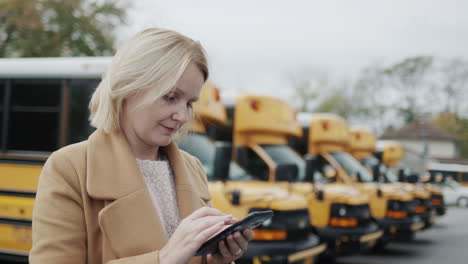 A teacher uses a smartphone, stands in the school yard against the background of yellow school buses. Side view
