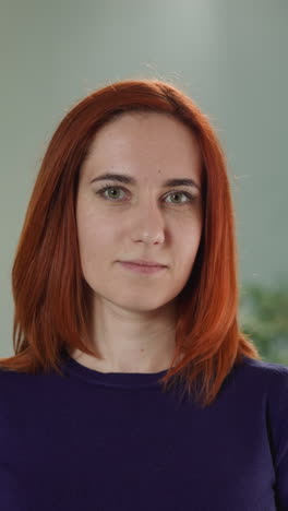 young redhead woman looks in camera slightly smiling. portrait of attractive female person standing against blurred lamp and pot-plant in room closeup