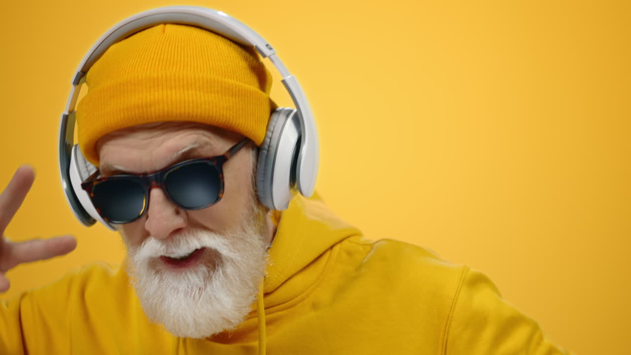 Joyful old man listening music in headphones indoors. Mature guy dancing inside.