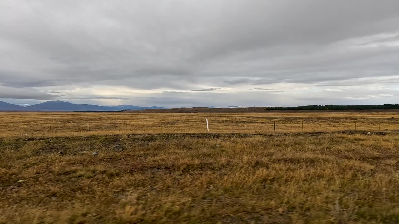 A moving vehicle travels beside a vast, grassy field under overcast skies, with distant mountains and roadside markers visible in a rural landscape