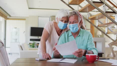 senior caucasian couple wearing face masks calculating finances at home