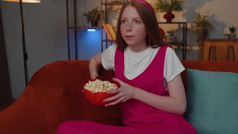 chica sentada en el sofá comiendo palomitas de maíz y viendo interesantes series de televisión, juegos deportivos en línea en casa