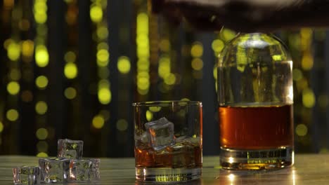 Barman-adds-ice-cubes-into-glass-with-golden-whiskey,-cognac-or-brandy-on-table.-Shiny-background