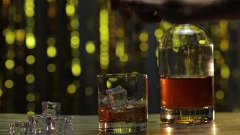 barman adds ice cubes into glass with golden whiskey, cognac or brandy on table. shiny background