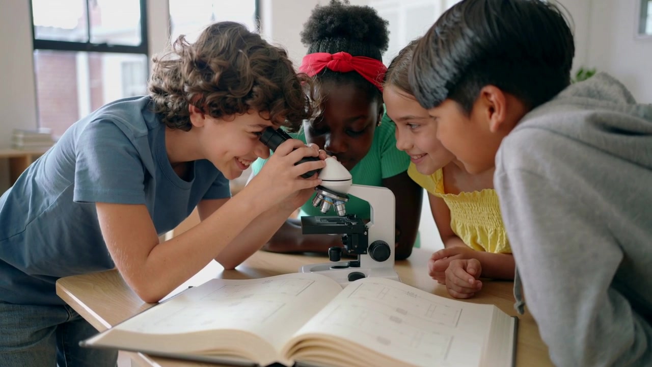 Positioning microscope, curly boy peering while peers leaning in class studying slide with book