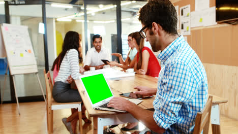 Businessman-using-laptop-and-digital-tablet-while-coworker-having-a-discussion-in-background