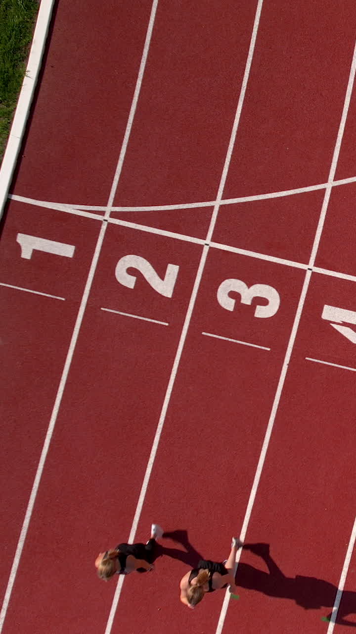 Vertical aerial shot of two young women sprinting on a red running track with lane numbers. Perfect for fitness, sports competition, and healthy lifestyle content