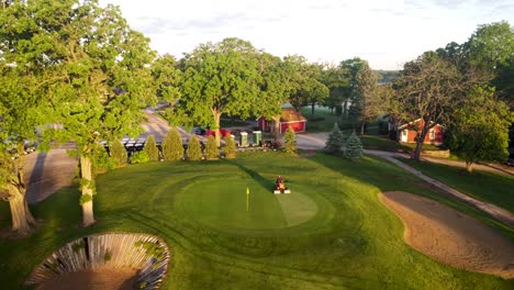 Vista-Aérea-De-Cortar-El-Césped-Alrededor-Del-Agujero-Con-Bandera-En-Un-Campo-De-Golf-Al-Atardecer