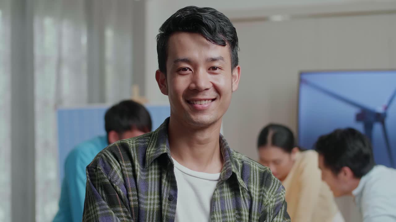 미소 짓는 아시아 남성 엔지니어가 동료들이 사무실에서 풍력 터빈에 대해 논의하는 동안 카메라에 팔을 고 있습니다.