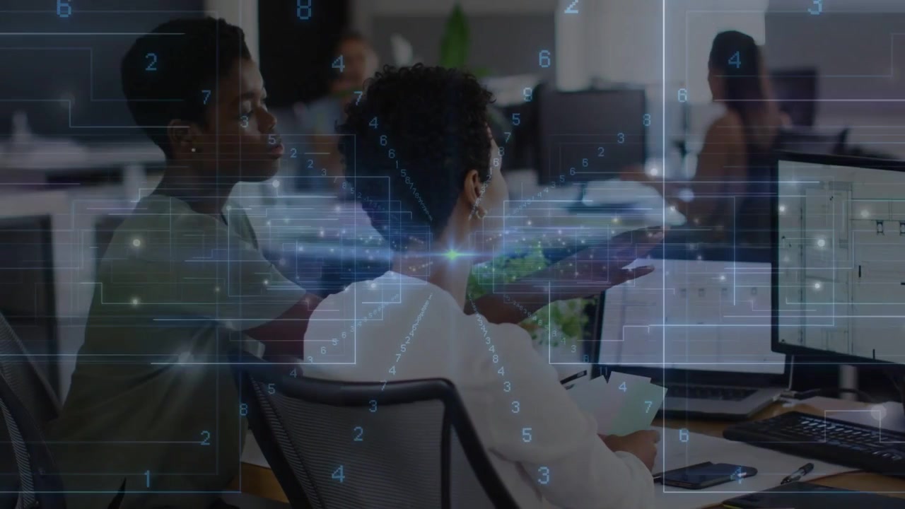 Seated worker using keyboard, peer leaning over and pointing, HUD showing monitors for data review