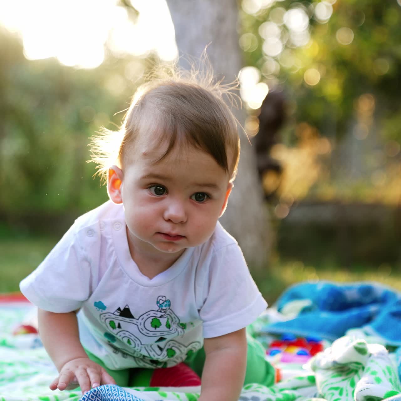 Niedlicher, kleiner Kleinkind, das über das im Garten auf dem Boden liegende Plaid krabbelt. Neugieriges, wunderschönes Kind, das sich interessiert umschaut. Verschwommener Hintergrund
