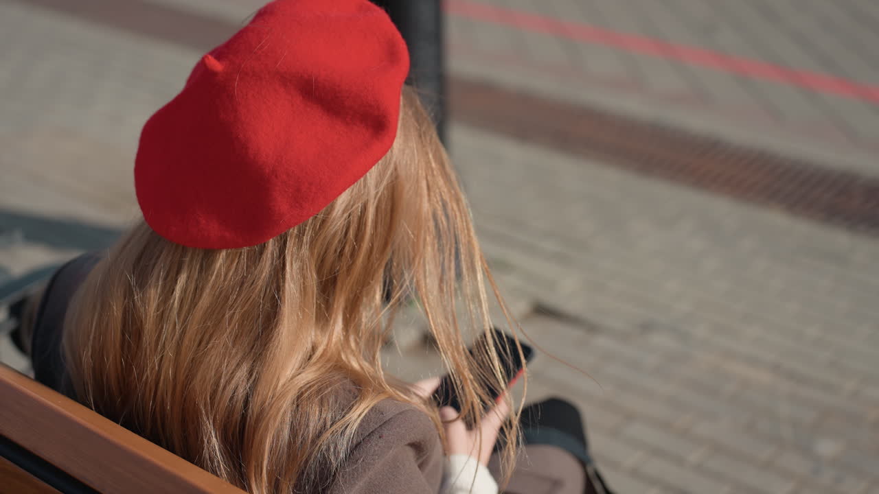 Hvid kvinde med rød baret scroller på telefon, nærbillede bagfra, blondt hår fanger sollys, modeblogger svarer på kommentarer, detalje med bænklampen, negle tapper på skærmen, personlig influencer.