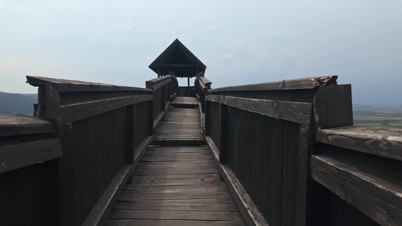Forward tracking shot of a wooden walkway leading to a covered observation deck, offering panoramic views of the scenic hills and fields in the tranquil countryside of Boldogkőváralja, Hungary