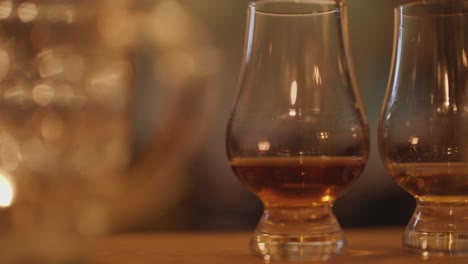 two whisky tasting glasses sit on a wooden table, surrounded by blurred glassware and soft candlelight, creating a cozy, intimate atmosphere