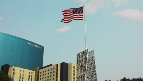 Bandera-Estadounidense-Ondeando-En-El-Asta-De-La-Bandera-Detrás-Del-Colorido-Edificio-Con-El-Hotel-Jw-Marriott-En-La-Distancia-En-Indianapolis,-Estados-Unidos