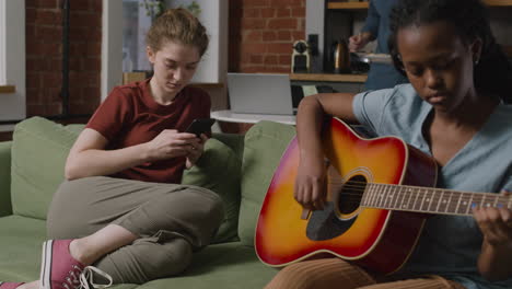 chica afroamericana tocando la guitarra sentada en el sofá mientras su compañera de cuarto usa el teléfono inteligente