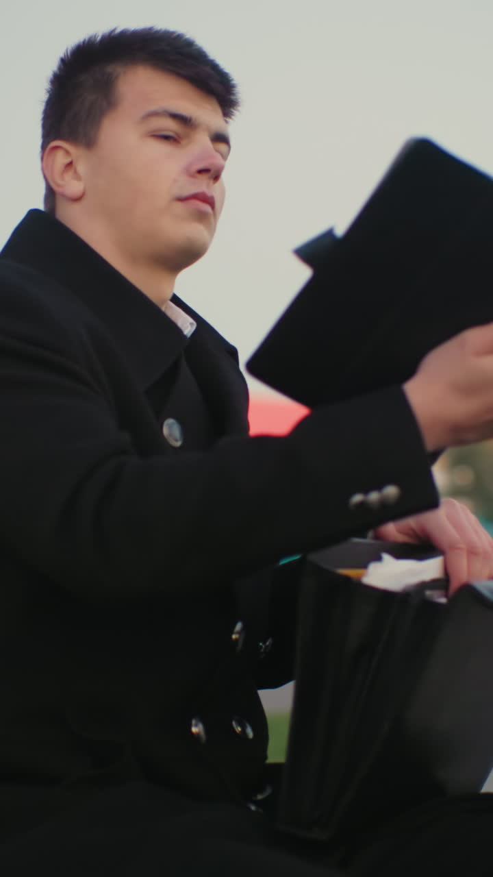 hombre sentado al aire libre revisando un maletín en busca de algo, sacando una tableta de la bolsa, con un fondo con edificios urbanos modernos y un entorno callejero de la ciudad