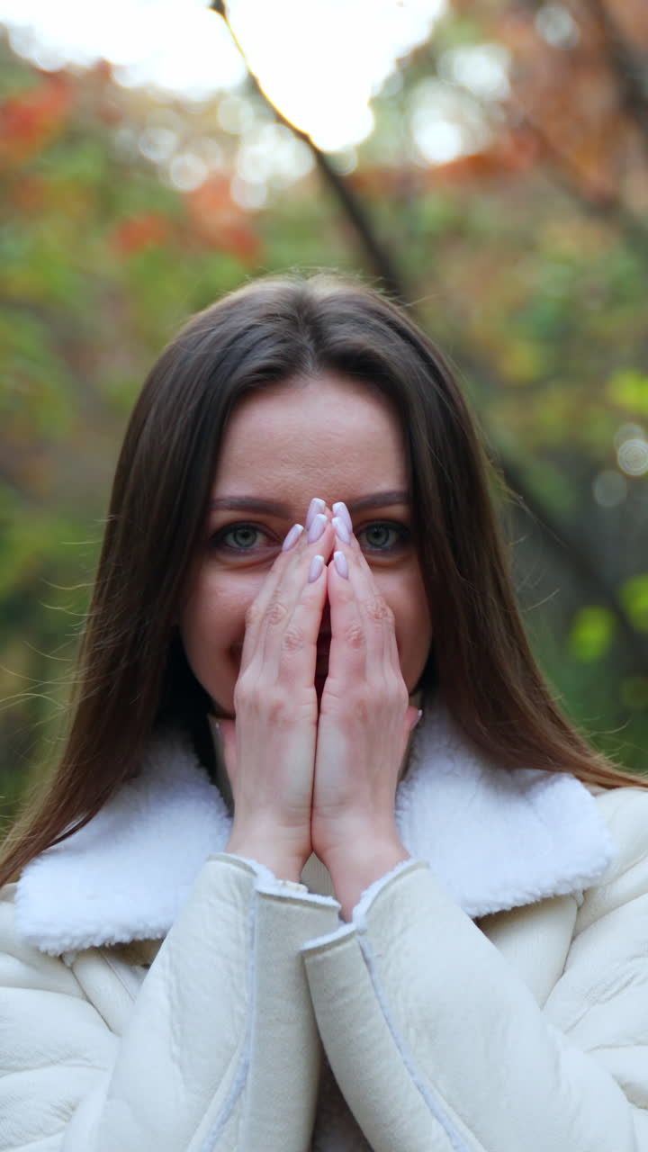 Beautiful rejoicing woman wearing white warm jacket taking a stroll by the forest. Lady warms her hands and enjoys fresh autumn air. Vertical video
