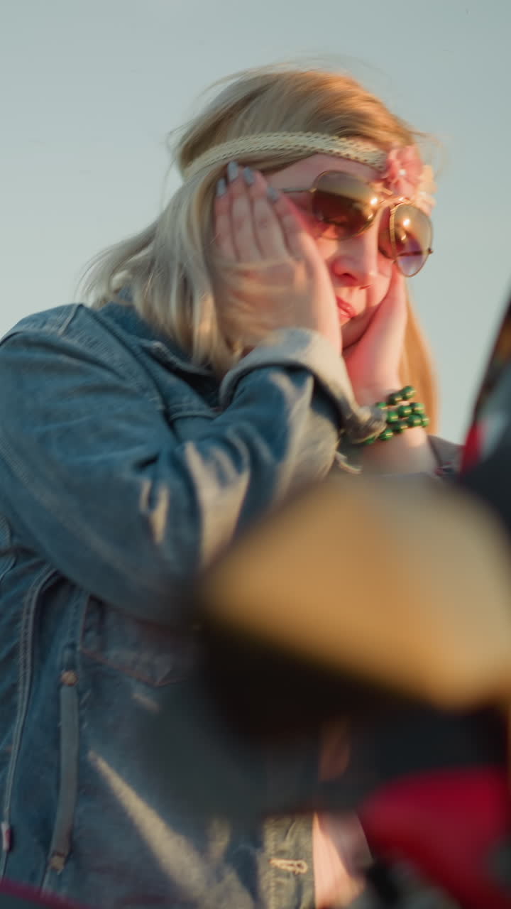 A woman wearing sunglasses and a flower headband is dancing with a joyful expression, moving her hands close to her face while standing near a red motorcycle in an open field