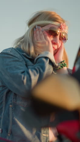 a woman wearing sunglasses and a flower headband is dancing with a joyful expression, moving her hands close to her face while standing near a red motorcycle in an open field