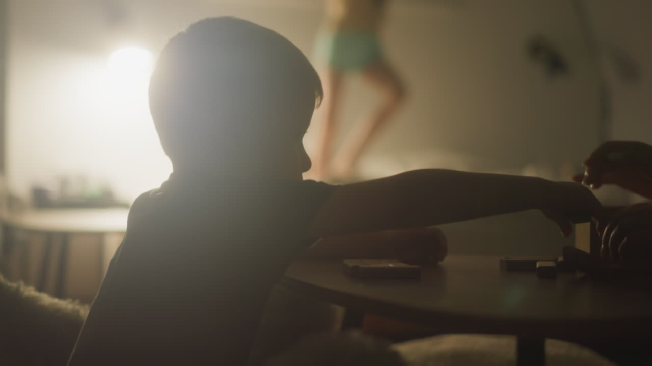 어린 소년은 작은 테이블에 앉아있는 동안 게임을 위해 나무 탑을 짓는 어머니를 도와줍니다. 아이는 여동생이 재미있고 침대에 뛰어다니는 배경에 앉습니다.