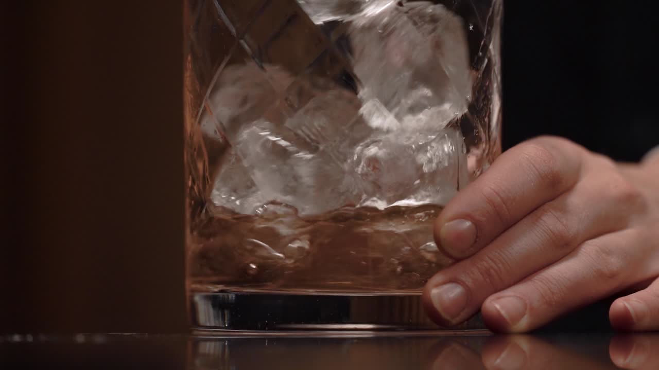A bartender is pouring spirit and liquor into the stirring glass and start to stir along a lot of ice using a long copper bar spoon.