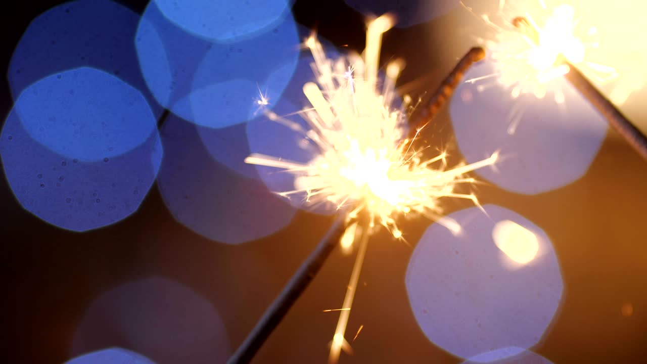 A close-up macro shot of two burning New Year sparklers. Bengal sparkler scattering into small sparkles against a background of blue garlands in blur. 4k slow motion