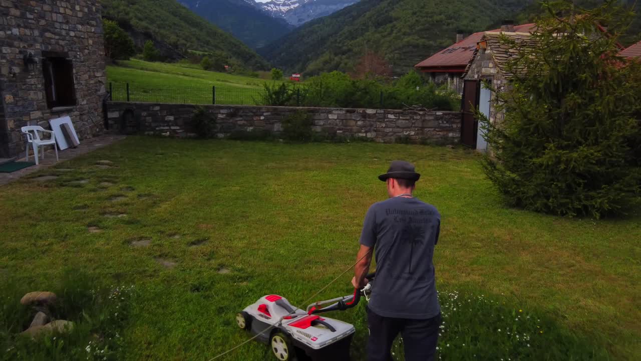 Aerial view of a man cutting the grass at home, with the electric grass cutting machine. In a village in the mountains.
