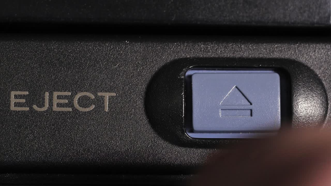 Finger presses blue eject key on right beside gold eject label on left of black camcorder