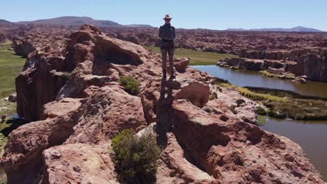 Aerial-follows-woman-climbing-rocky-bluff-above-valley-lake-in-Bolivia