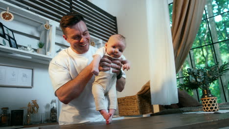 muscular caucasian man holds in a hands his tiny son. daddy puts an infant on the wooden table so the kid's feet touch the surface.
