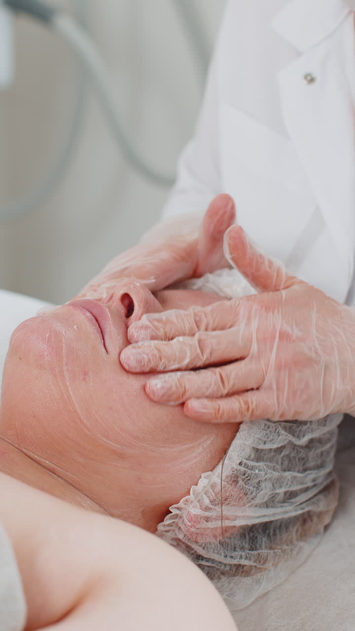 Cosmetologa applica una speciale crema idratante, scrub o peeling sul viso di una graziosa donna caucasica.
