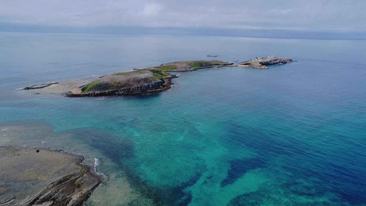 ブラジルのアブロリョス諸島。透明なターコイズブルーと深い青色の大西洋の海に浮かぶ岩の島々が広がる空撮風景を、ドローンが前方に滑空しながら捉える。